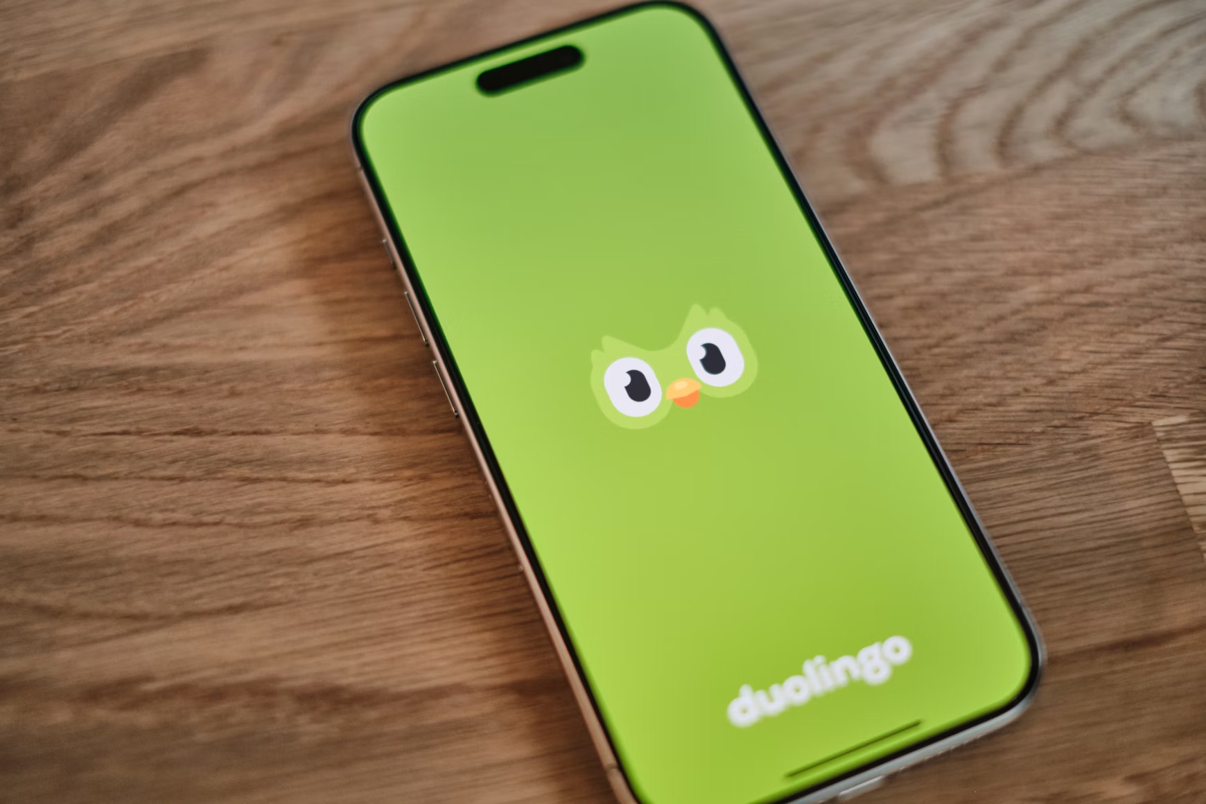 iPhone on a wooden surface showing Duolingo's green app launch screen with the owl mascot