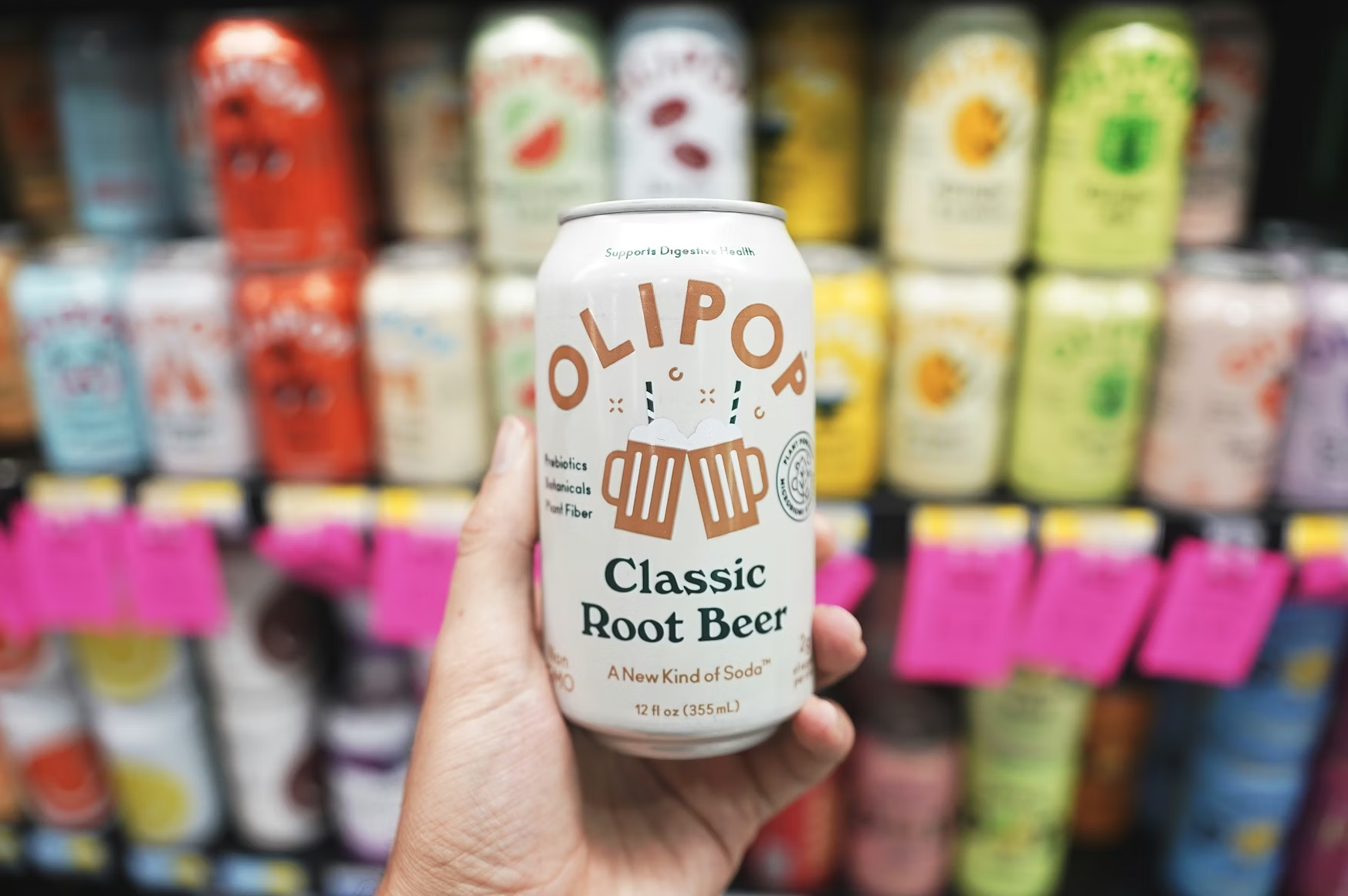 Hand holding an Olipop Classic Root Beer can in front of a colorful wall of Olipop flavor variants
