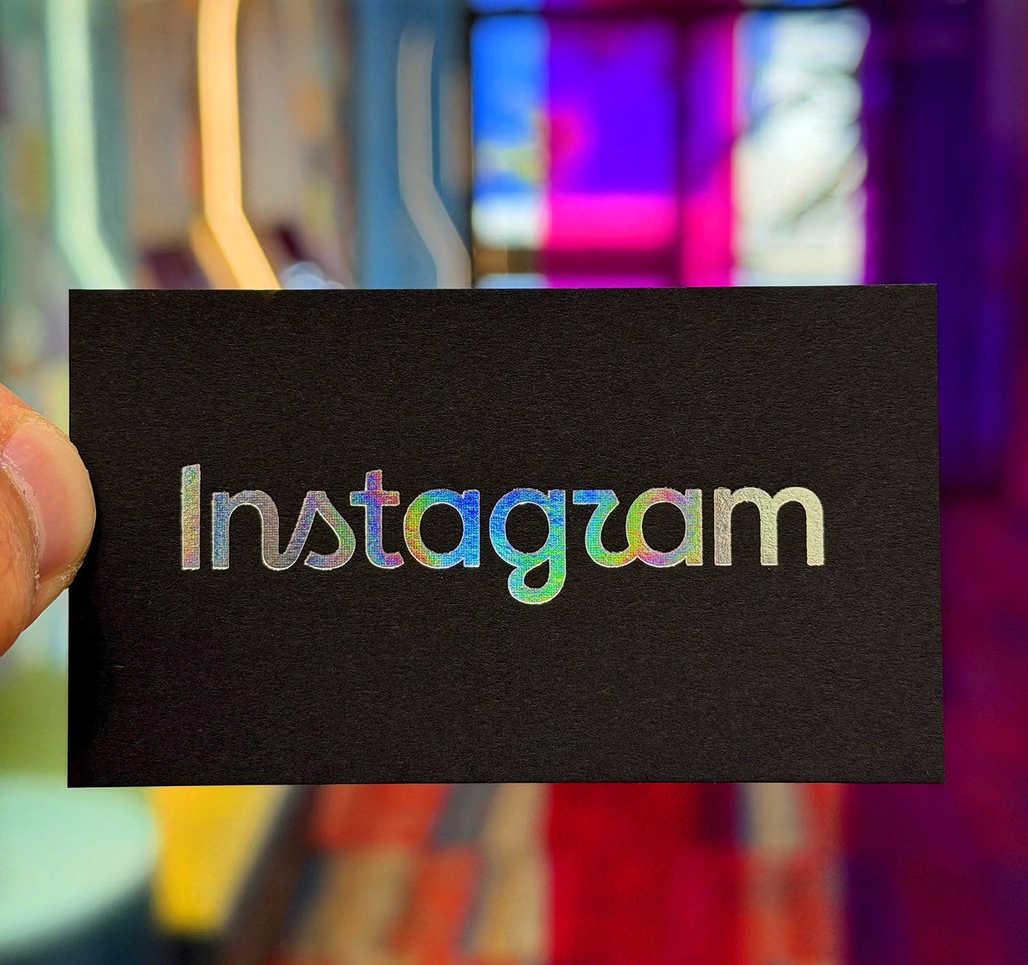 Instagram's 2026 wordmark stamped in holographic foil on a black business card, held up to the light so the foil shifts through blue, green and pink across the letterforms